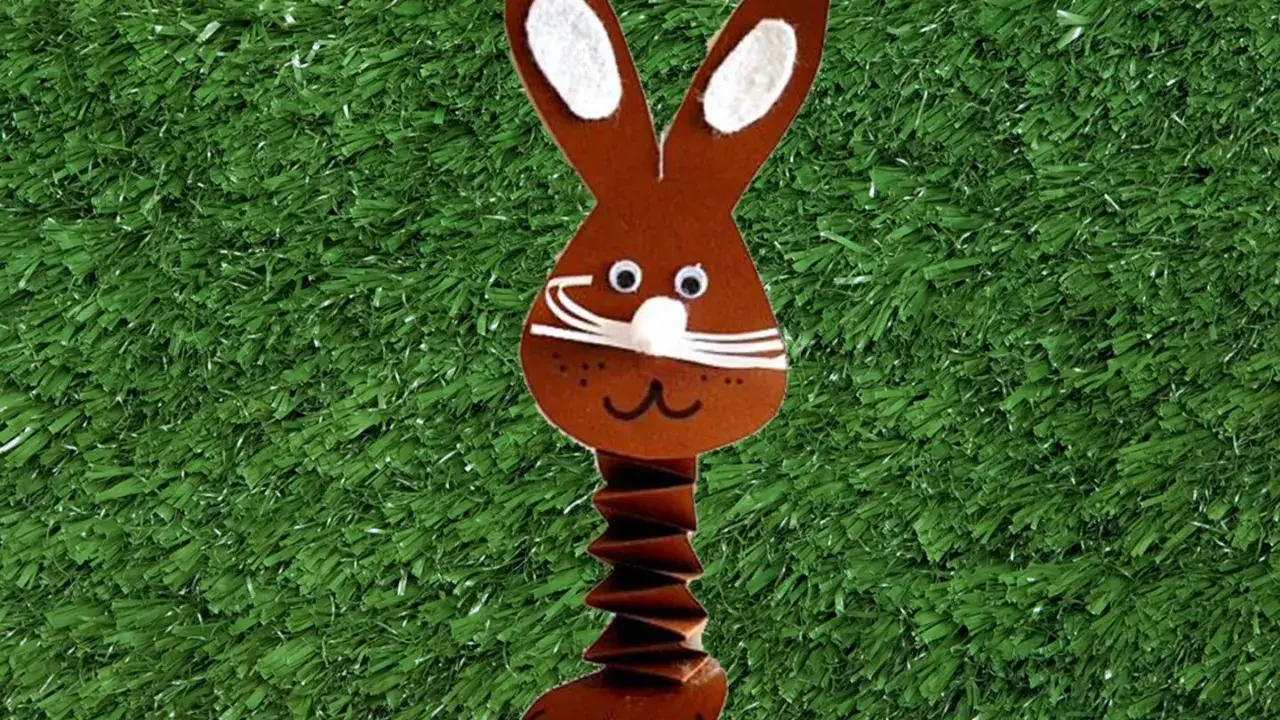 Brauner Papierhase mit Ziehharmonika-Körper, ein süßes Beispiel für Osterhasen basteln, auf grünem Kunstrasen mit bunten Eiern.