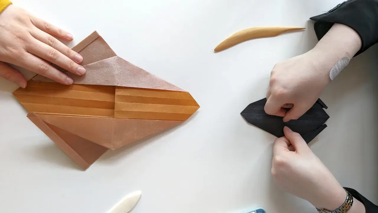 Zwei Hände falten Papier, eine an einem komplexen braunen Origami, die andere an einem schwarzen. Eine schwierige Origami-Anleitung wird befolgt.