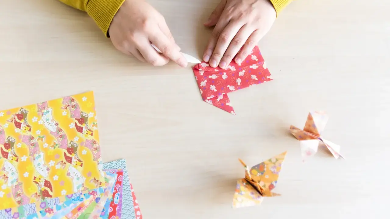 Hände falten Papier für eine Origami Kugel Anleitung. Mehrere bunte Origami-Papierbögen liegen bereit.