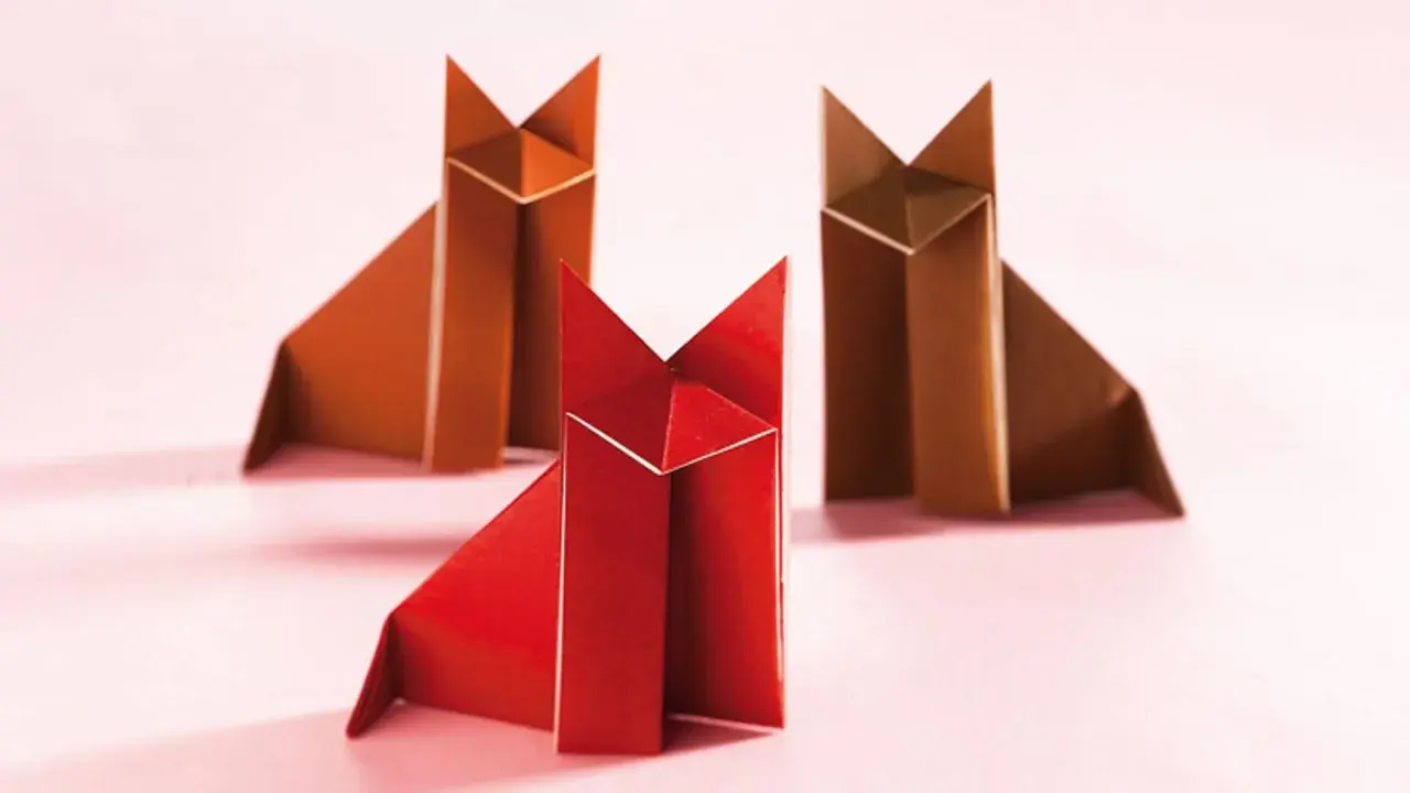 Drei origami Füchse, einer rot und zwei braun, sitzen auf einem rosa Untergrund.