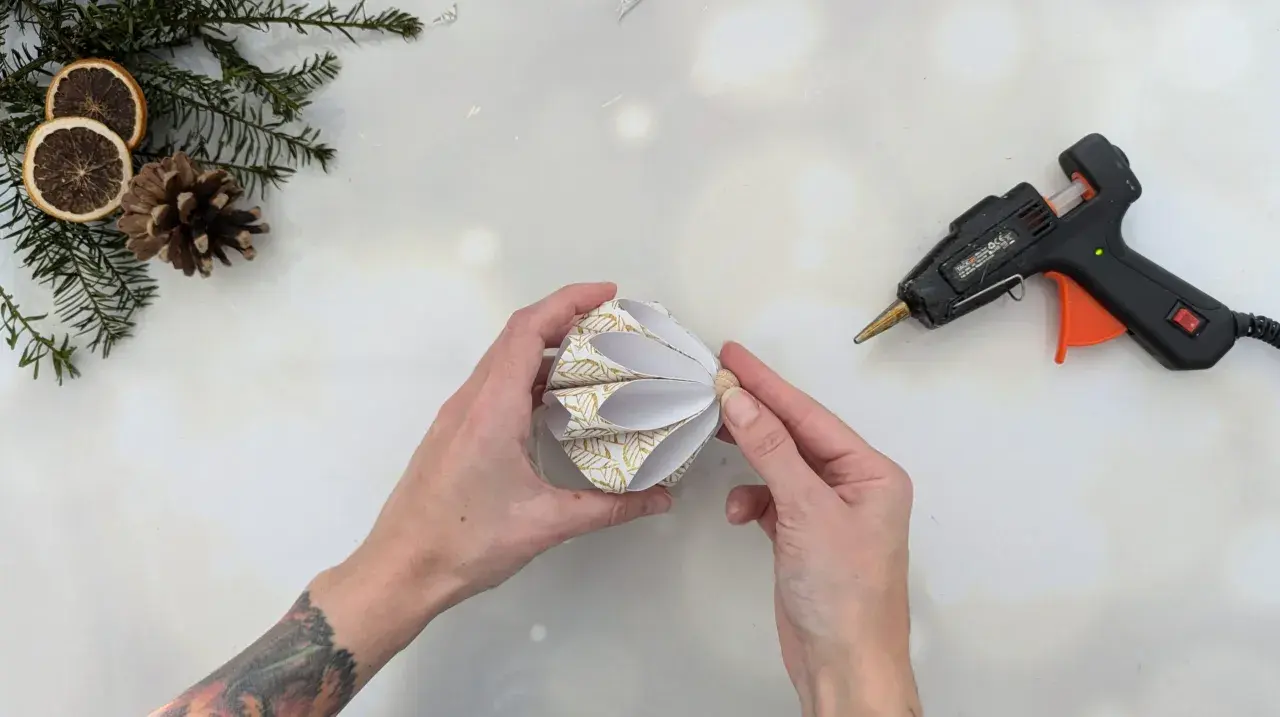 Hände basteln mit Klebepistole und Papier einen schönen DIY Christbaumschmuck. Tannenzweige und Orangenscheiben liegen bereit.