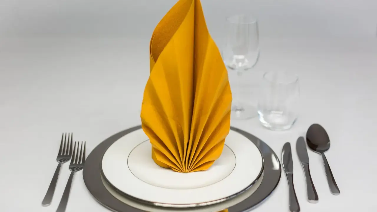 Ein festlich gedeckter Tisch mit einem gelben, gefalteten Servietten-Fächer. Gedeck mit Tellern, Besteck und Gläsern.