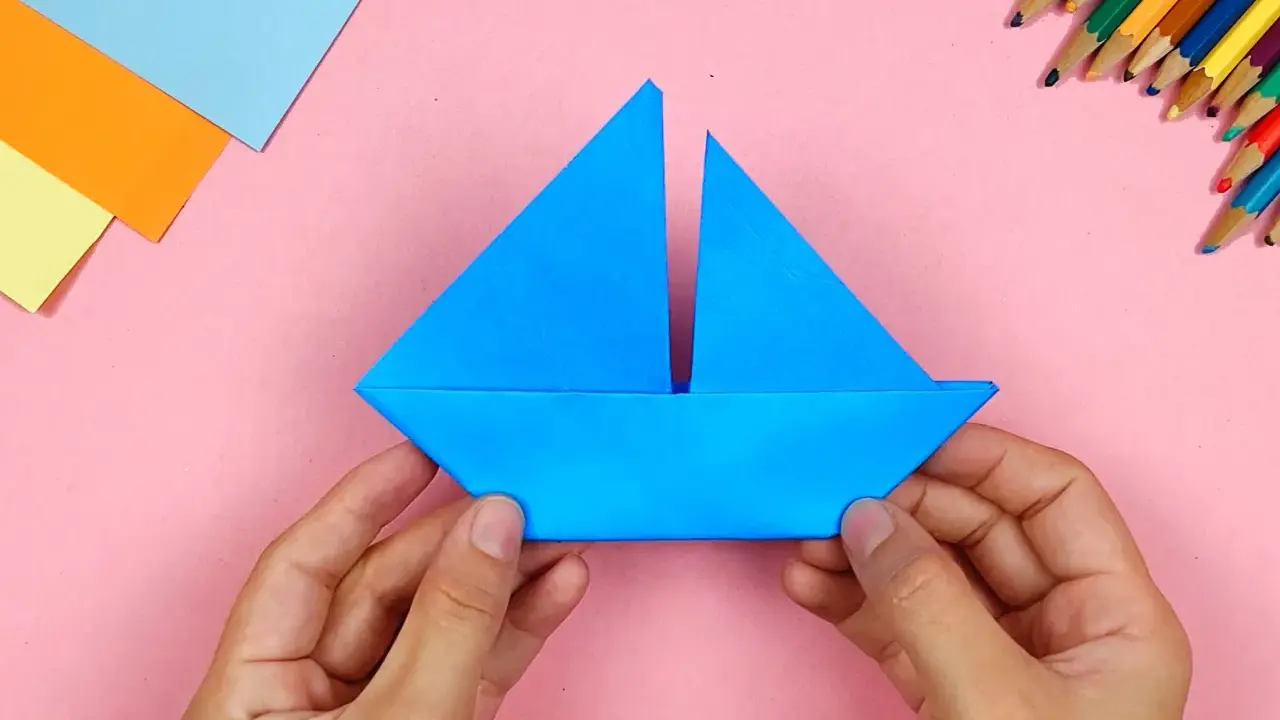 Hände falten ein blaues Papierboot. Bunte Stifte und Papier liegen bereit für weitere Bastelprojekte.