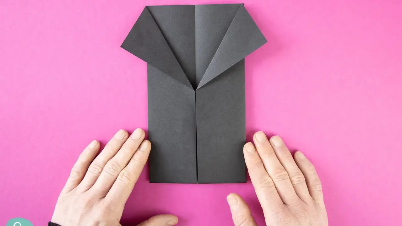 Ein schwarzes Origami-Hemd wird auf pinkem Hintergrund gefaltet. Hände halten das Papier.