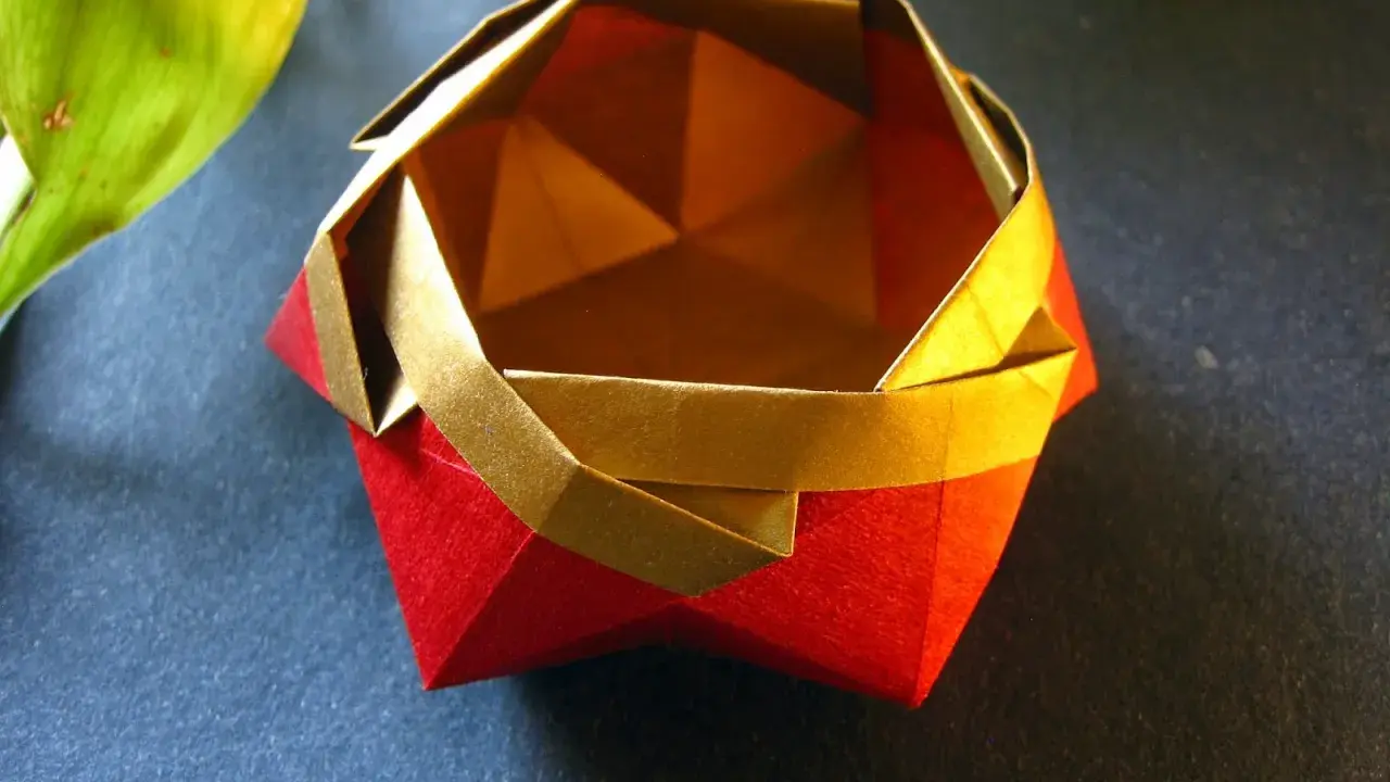 Eine kunstvolle origami schale in Rot und Gold, daneben ein grünes Blatt.