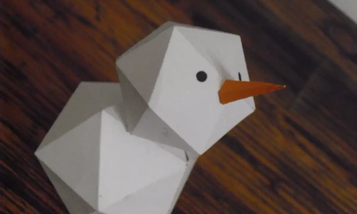 Geometrischer Schneemann, gefaltet aus Papier, mit orangefarbener Karotte als Nase und schwarzen Augen.