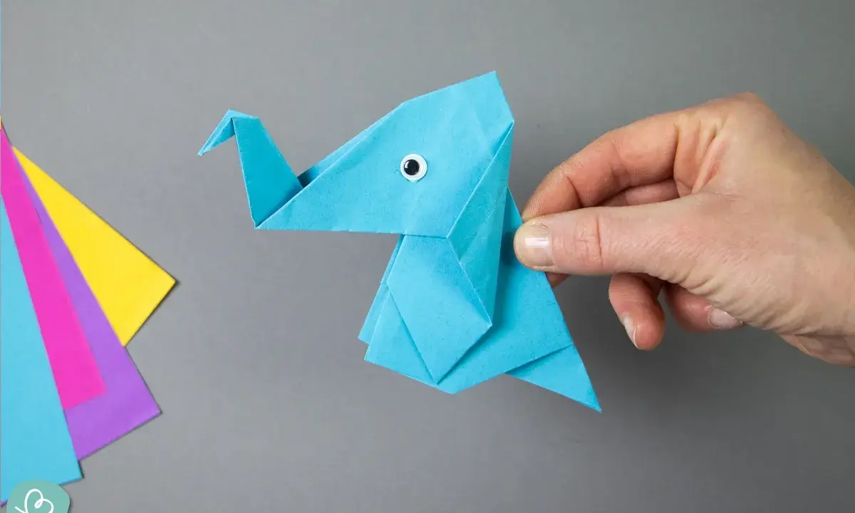 Ein blauer origami elefant mit einem kleinen Knopfauge wird von einer Hand gehalten. Daneben liegen bunte Papierbögen.
