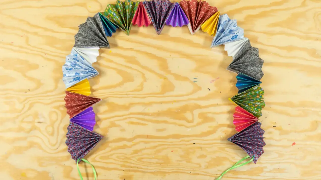 Bunte Papierfächer, die zu einer ziehharmonika-girlande gebastelt wurden, liegen auf einer Holzplatte.