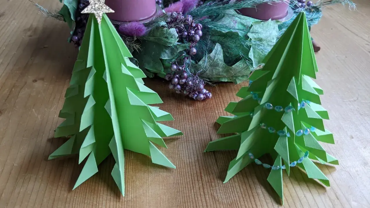 Zwei grüne, flache Origami Tannenbäume stehen auf einem Holztisch. Einer ist mit einer goldenen Sternspitze geschmückt, der andere mit einer Perlenkette. Im Hintergrund ein Adventskranz.