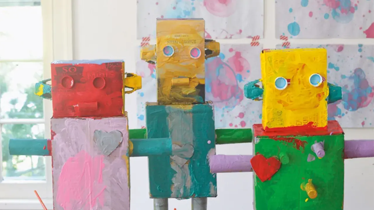 Drei bunte Roboter, die Kinder gebastelt haben, stehen nebeneinander. Sie sind aus Kartons und Rollen gefertigt und mit leuchtenden Farben bemalt.