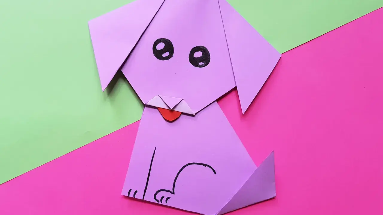 Ein lila **Falten Hund** sitzt auf pinkem und grünem Papier. Er hat schwarze Augen und eine rote Zunge.