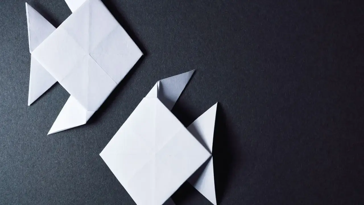 Zwei weiße Origami-Fische, kunstvoll gefaltet, schwimmen auf dunklem Grund.
