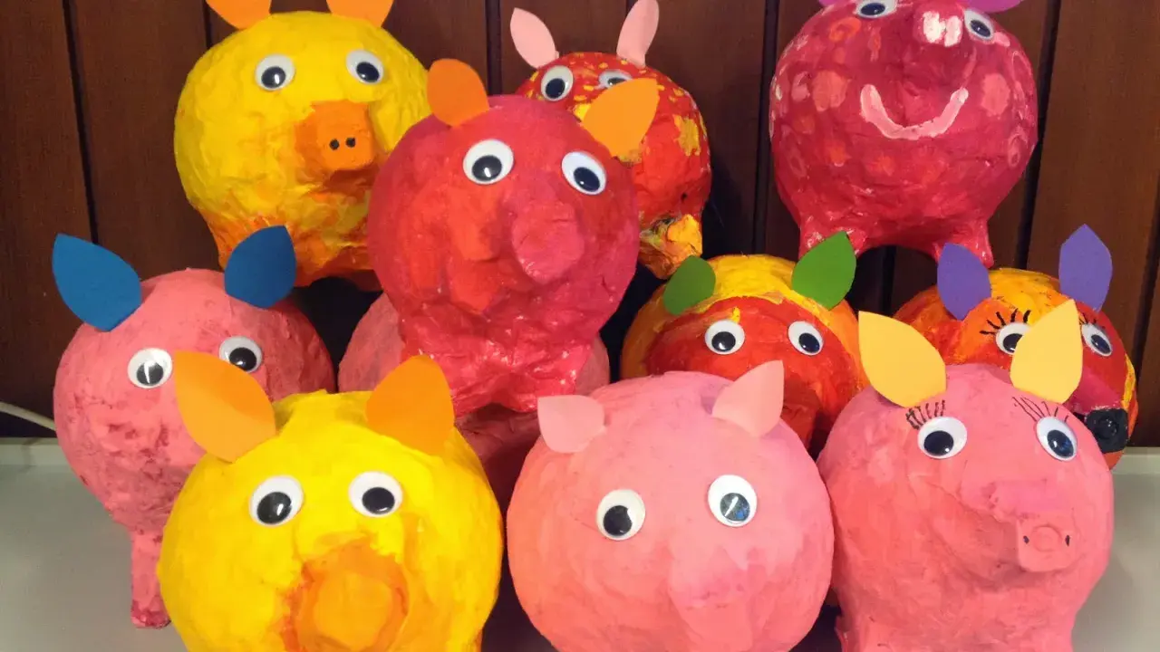 Bunte Pappmaché-Sparschweine mit großen Kulleraugen und Papierohren stehen fröhlich nebeneinander.