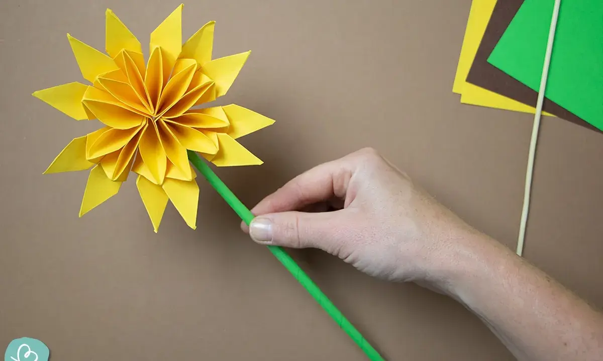 Eine Hand hält eine gelbe origami sonne mit einem grünen Stiel. Bastelmaterialien liegen daneben.