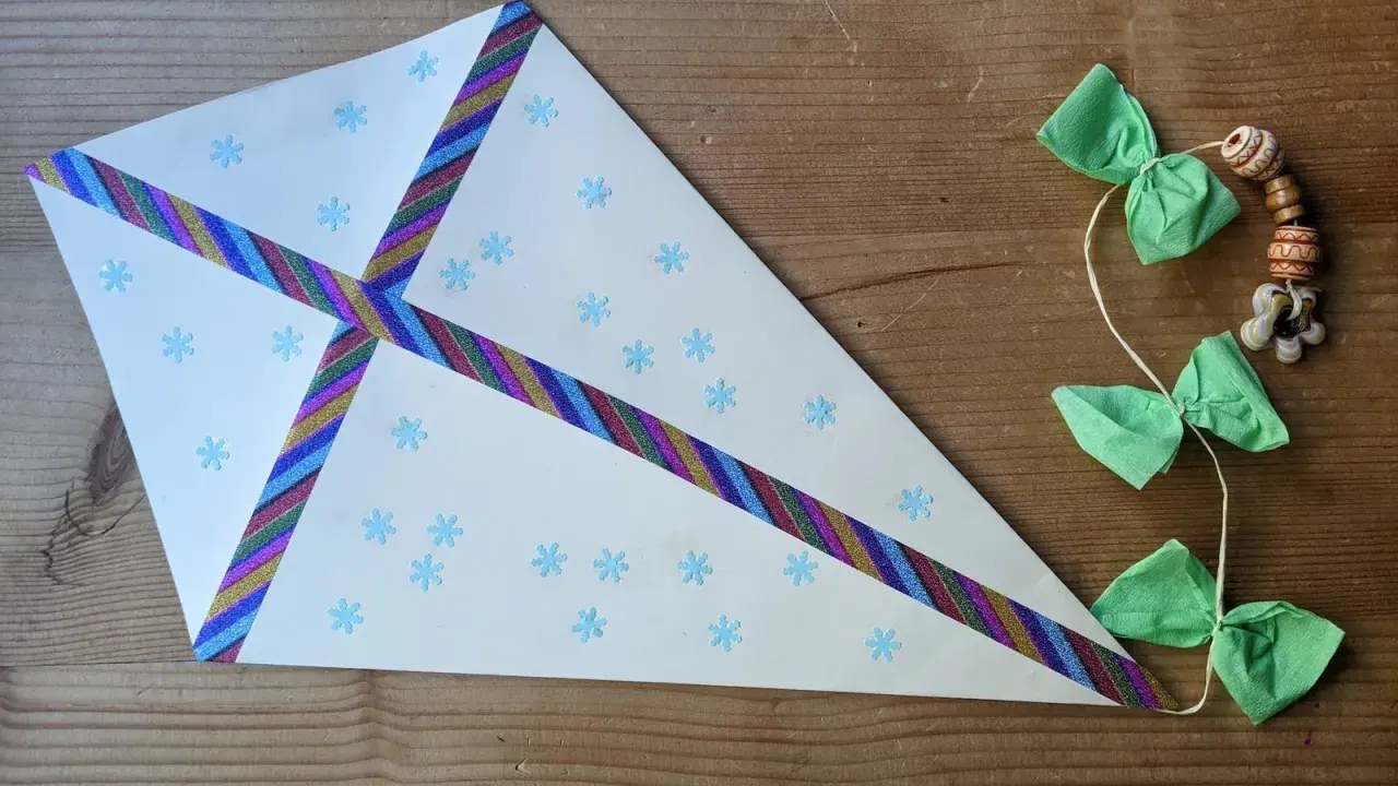 Bastelanleitung mit Bildern: Ein selbstgebastelter Drachen aus Papier mit Schneeflocken und buntem Klebeband, geschmückt mit grünen Schleifen und Holzperlen.