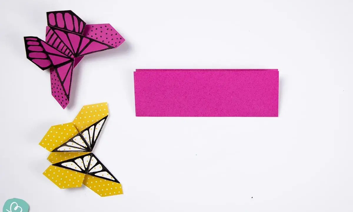 Zwei bunte origami schmetterlinge, einer pink mit Punkten, der andere gelb mit Punkten und Flügelmustern, liegen neben einem Streifen pinken Papiers.