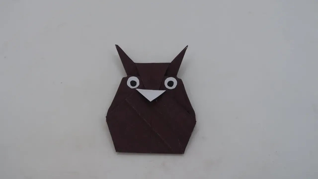 Eine braune origami Eule mit großen weißen Augen und einem kleinen Schnabel sitzt auf hellem Untergrund.