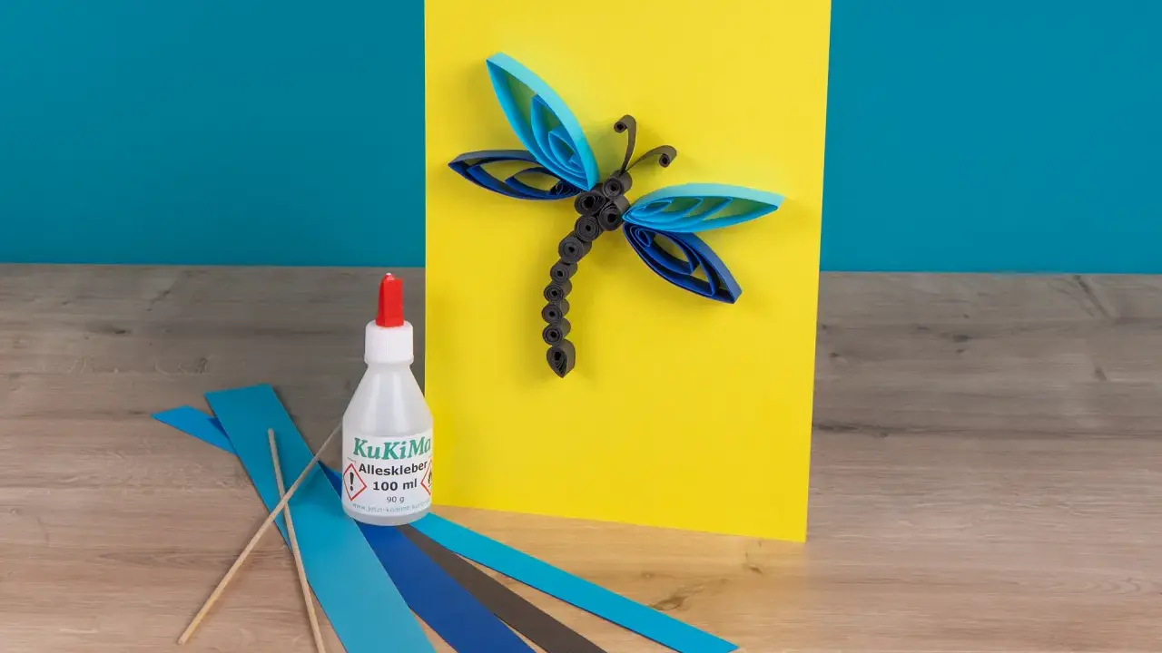Eine Libelle aus Papierstreifen, erstellt mit einer Quilling-Anleitung. Kleber und bunte Streifen liegen bereit.