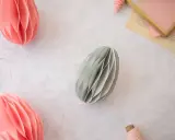 Ostereier basteln Papier: Dekorative Papier-Ostereier in Rosa und Grau hängen an Schnüren. Ein Garnknäuel und eine Schere liegen daneben.
