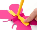 Hand bastelt fliegende Schmetterlinge aus pinkem Papier. Ein gelber Streifen wird befestigt.
