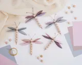 Vier selbstgebastelte Libellen aus Holzperlen und Papierflügeln, inspiriert von einer libelle basteln vorlage. Zarte Pastellfarben und ein Musselintuch schaffen eine frühlingshafte Atmosphäre.