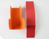 Zwei Bastelbögen, einer orange, der andere rot, liegen nebeneinander. Der rote Bogen ist gefaltet, bereit, um daraus Stiefel zu falten.