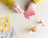 Hände falten Papier für eine Origami Kugel Anleitung. Mehrere bunte Origami-Papierbögen liegen bereit.