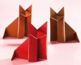 Drei origami Füchse, einer rot und zwei braun, sitzen auf einem rosa Untergrund.