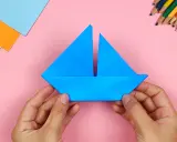 Hände falten ein blaues Papierboot. Bunte Stifte und Papier liegen bereit für weitere Bastelprojekte.