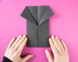 Ein schwarzes Origami-Hemd wird auf pinkem Hintergrund gefaltet. Hände halten das Papier.