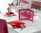 Eine 3D-Karte basteln zum Geburtstag: Rote Herzen auf einer Klappkarte, Stempel, Federn und Stifte liegen bereit.