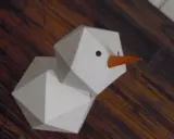 Geometrischer Schneemann, gefaltet aus Papier, mit orangefarbener Karotte als Nase und schwarzen Augen.