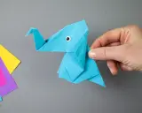 Ein blauer origami elefant mit einem kleinen Knopfauge wird von einer Hand gehalten. Daneben liegen bunte Papierbögen.