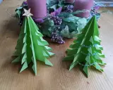 Zwei grüne, flache Origami Tannenbäume stehen auf einem Holztisch. Einer ist mit einer goldenen Sternspitze geschmückt, der andere mit einer Perlenkette. Im Hintergrund ein Adventskranz.