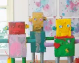Drei bunte Roboter, die Kinder gebastelt haben, stehen nebeneinander. Sie sind aus Kartons und Rollen gefertigt und mit leuchtenden Farben bemalt.