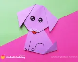 Ein lila **Falten Hund** sitzt auf pinkem und grünem Papier. Er hat schwarze Augen und eine rote Zunge.