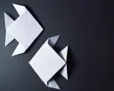 Zwei weiße Origami-Fische, kunstvoll gefaltet, schwimmen auf dunklem Grund.