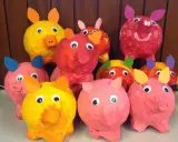 Bunte Pappmaché-Sparschweine mit großen Kulleraugen und Papierohren stehen fröhlich nebeneinander.