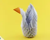 Ein weißer Schwan aus modularem Origami mit orangefarbenem Schnabel steht vor einem gelben Hintergrund.
