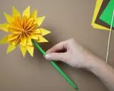 Eine Hand hält eine gelbe origami sonne mit einem grünen Stiel. Bastelmaterialien liegen daneben.