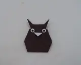 Eine braune origami Eule mit großen weißen Augen und einem kleinen Schnabel sitzt auf hellem Untergrund.