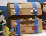 Bastelidee: Eine Schatztruhe aus Pappe, verziert mit Piratenmotiven und gefüllt mit Goldmünzen. Ein Affe hält eine Piratenflagge.
