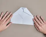 Hände falten einen weißen Papierflieger. Der runder Papierflieger nimmt langsam Form an.