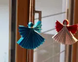 Zwei glitzernde Origami Weihnachts Engel, einer in Blau, der andere in Rot und Silber, hängen an Fäden.