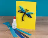 Eine Libelle aus Papierstreifen, erstellt mit einer Quilling-Anleitung. Kleber und bunte Streifen liegen bereit.