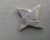 Ein silberner Papier-Wurfstern, perfekt zum **Wurfstern basteln**. Liegt auf einer texturierten, hellgrünen Unterlage.