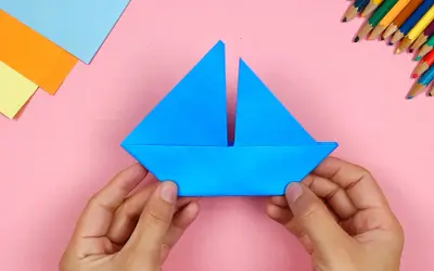 Hände falten ein blaues Papierboot. Bunte Stifte und Papier liegen bereit für weitere Bastelprojekte.