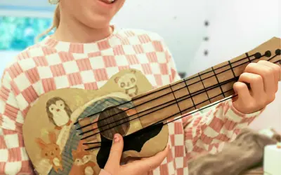 Kind bastelt Gitarre nach Vorlage. Die selbstgemachte Gitarre ist mit süßen Tieren wie Pinguin, Panda, Hase und Fuchs bemalt.