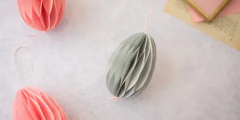 Ostereier basteln Papier: Dekorative Papier-Ostereier in Rosa und Grau hängen an Schnüren. Ein Garnknäuel und eine Schere liegen daneben.