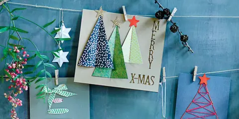Selbstgebastelte Weihnachtskarten mit Bäumen und Sternen hängen an einer Schnur. Ein grüner Zweig mit Beeren und Papiersterne zieren die Szene.